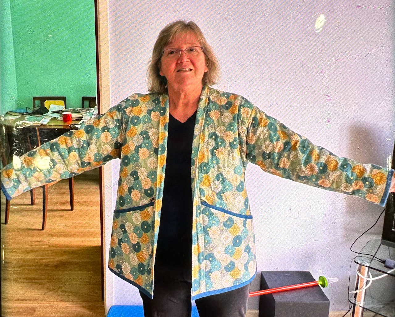 Customer wearing a handmade long jacket in sage Flowerella linen fabric, featuring colourful floral motifs and contrasting blue trim.