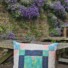 Valley Falls Quilt Block on a garden bench among small purple flowers. Pastel colours on the outside. Floral pattern inside.