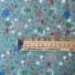 My Little Garden cotton fabric in pistachio, featuring a scattered floral pattern with white, coral, red, and blue blooms, taupe flowers, and green foliage on a sage green background. With ruler to show size of design.
